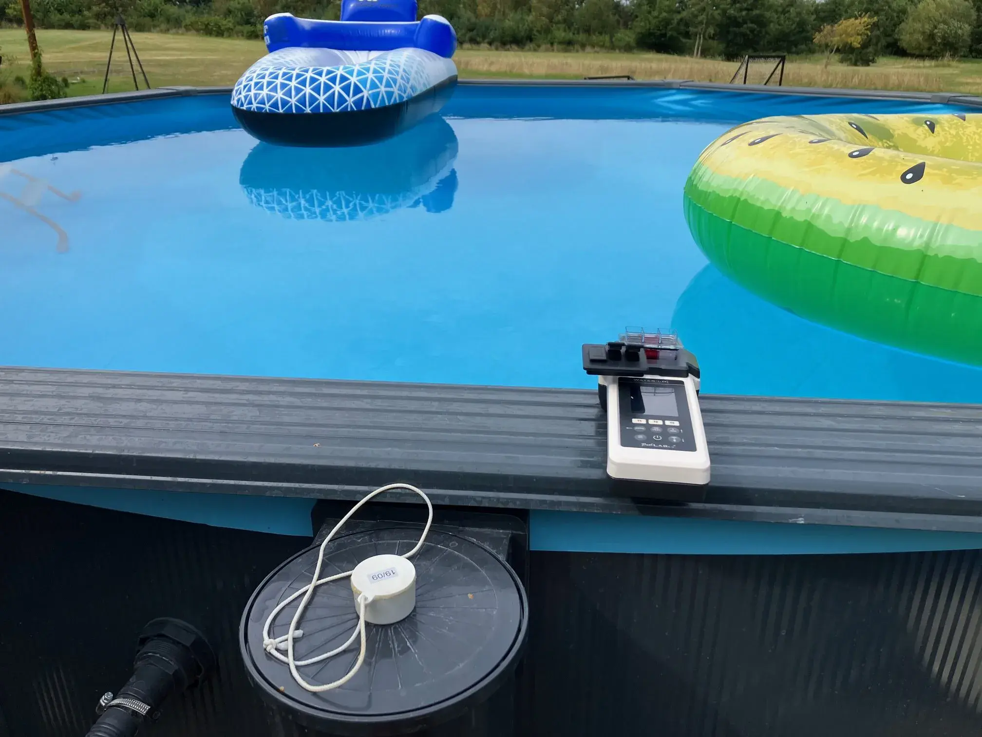 PoolLab-måler ligger på kanten af en pool ved siden af filterudstyr.